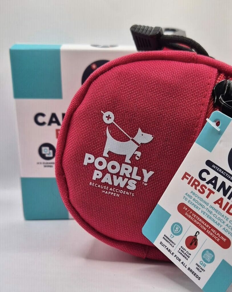 A bright pink "Poorly Paws" pet first aid kit with a white dog logo sits in front of a blue and white box. The kit has a tag indicating it's suitable for all breeds and includes emergency supplies for pets.