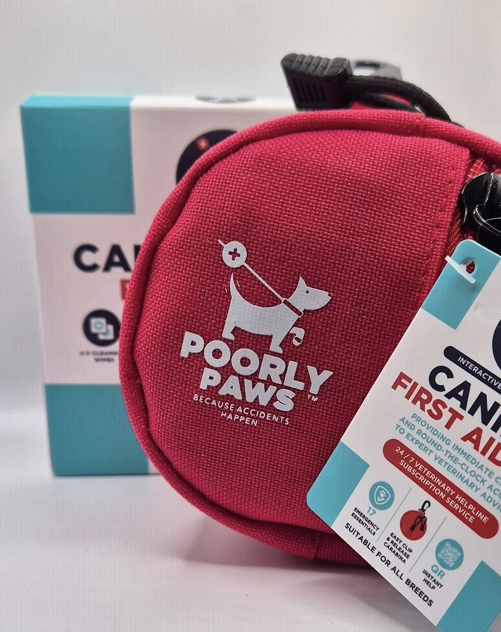 A bright pink "Poorly Paws" pet first aid kit with a white dog logo sits in front of a blue and white box. The kit has a tag indicating it's suitable for all breeds and includes emergency supplies for pets.