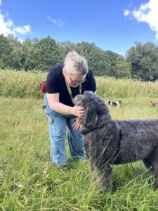 A person with blonde hair in a ponytail and glasses bends down to pet a large, fluffy black dog in a grassy field, with trees and a blue sky in the background.